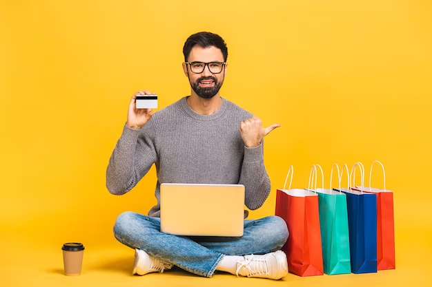 A Happy Customer Buying on a Computer A photo of a man sitting in front of a computer, holding up a credit card with shopping bags next to him.